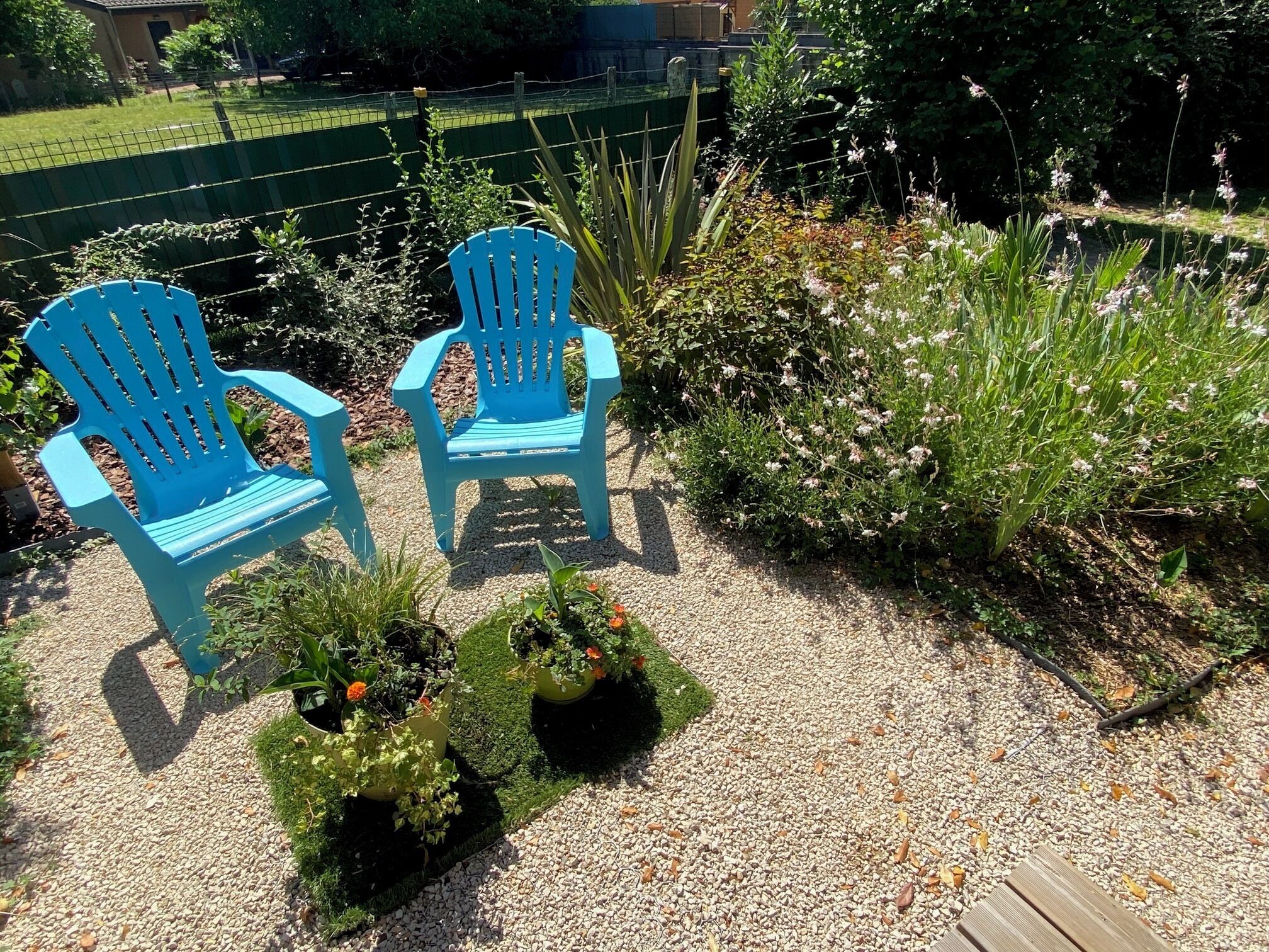 Two blue plastic chairs placed on a gravel path with potted plants nearby.