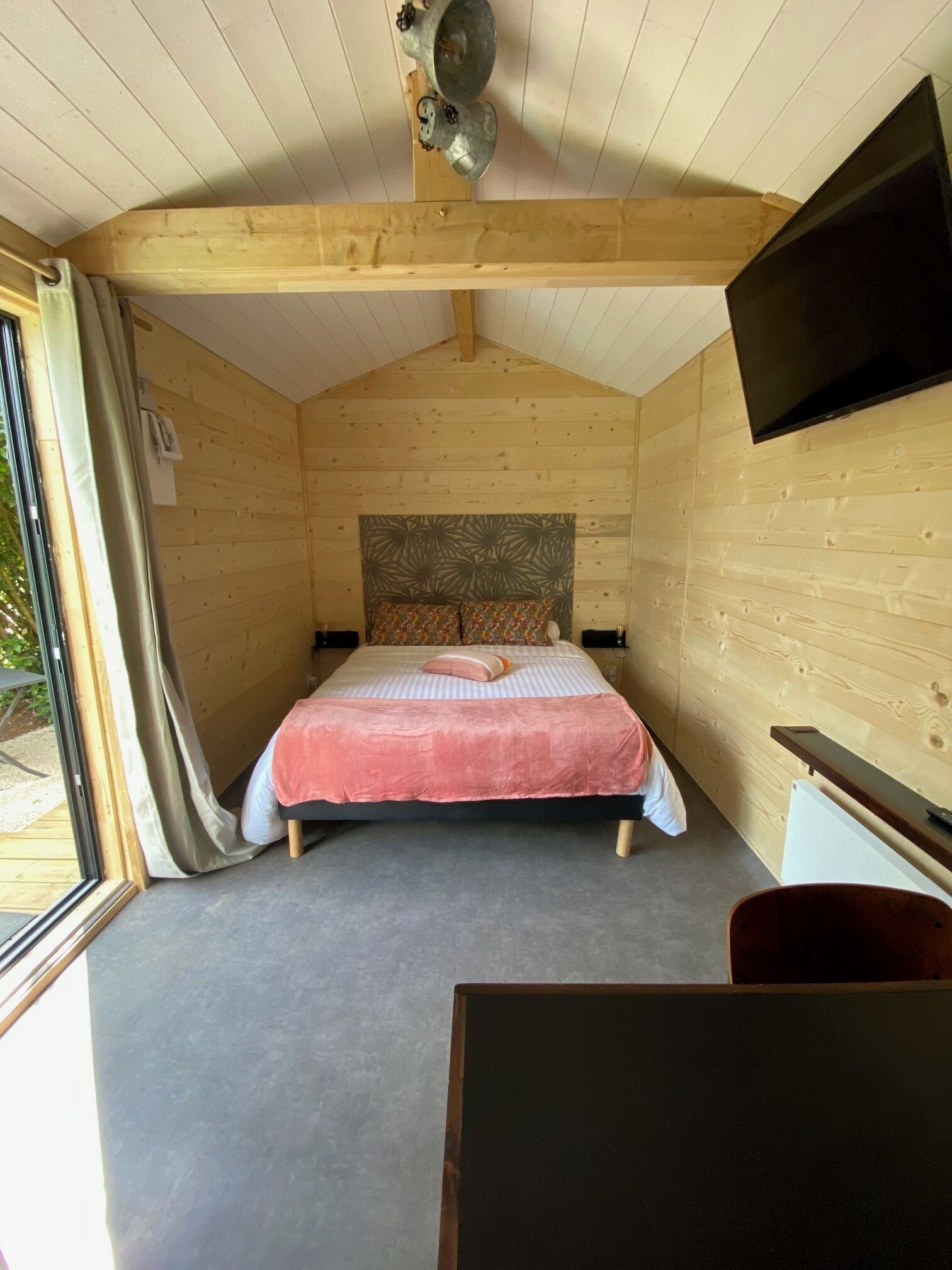 Une chambre confortable, aménagée dans une cabane en bois, avec un lit, un téléviseur fixé au mur et une fenêtre avec des rideaux.