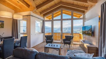 Ein gemütliches Wohnzimmer mit einer Couch, Sesseln, einem Couchtisch und einem an der Wand befestigten Fernseher. Große Fenster bieten Blick auf die Berge.