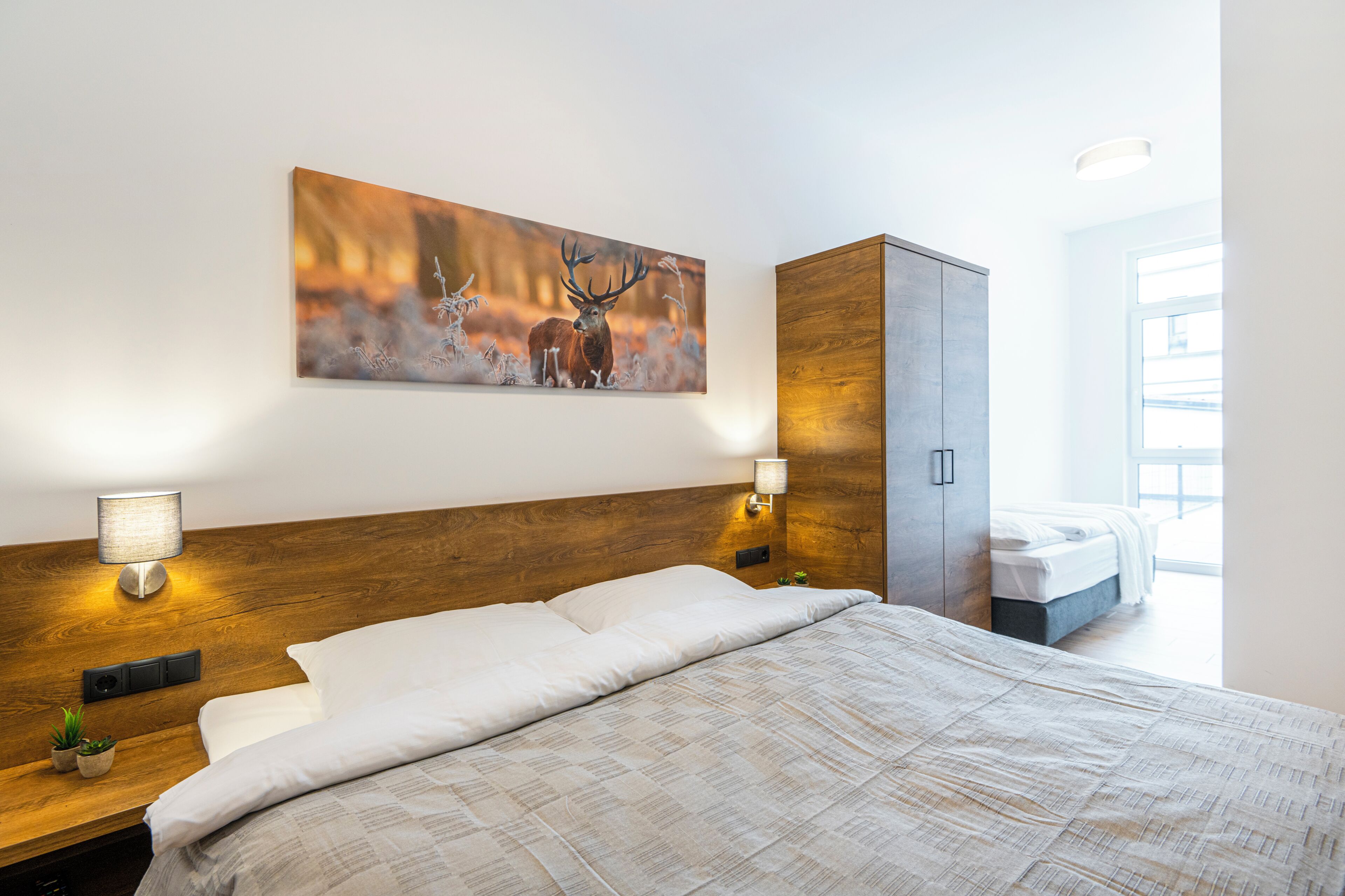 Habitación de hotel con una cama grande, cabecera de madera, mesita de noche con lámpara, armario y un cuadro de ciervos en la pared.