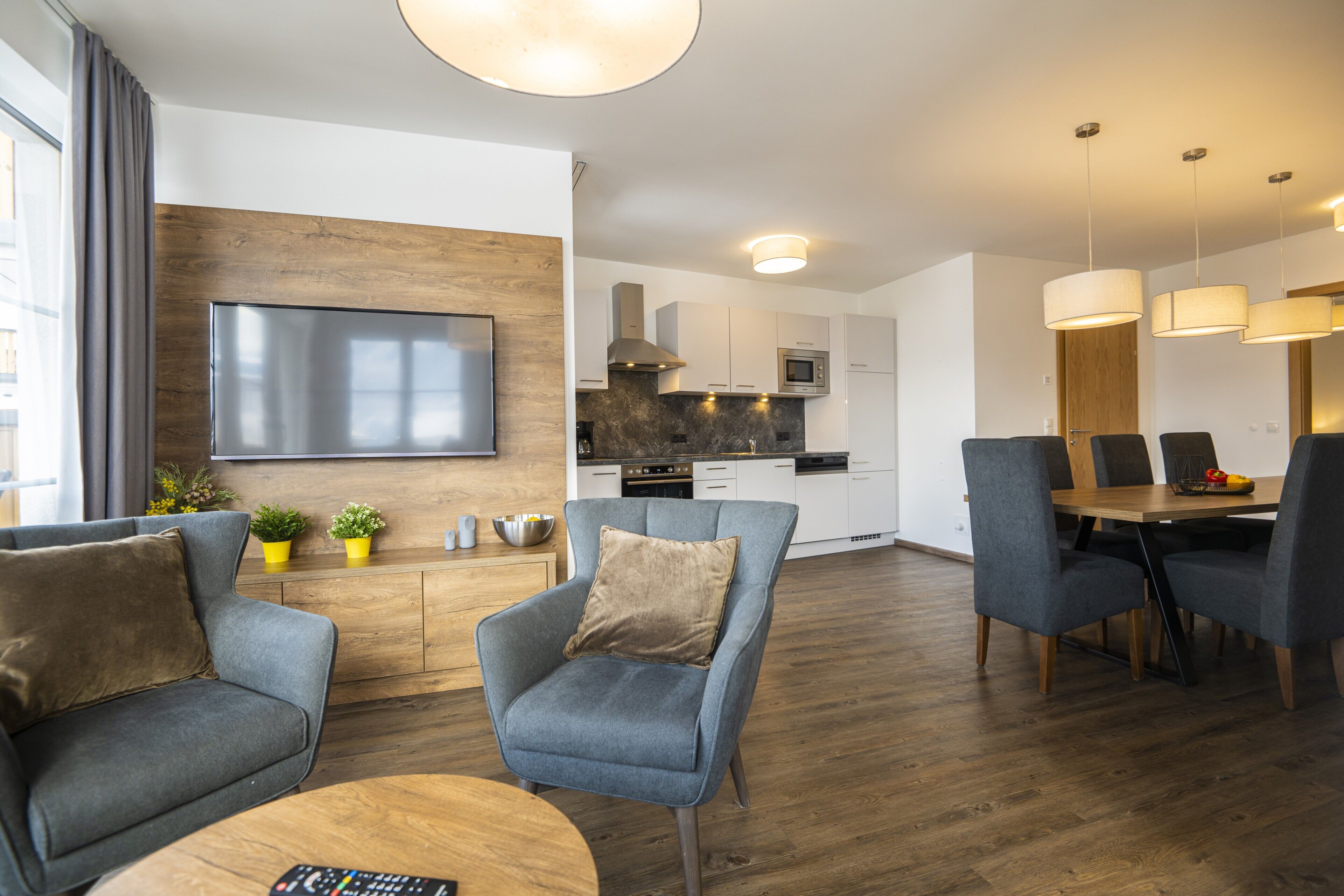 Una sala moderna con televisor de pantalla plana, sofás grises, una mesa de centro de madera y un comedor con mesa y sillas de madera.