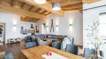 A modern living space with a wooden ceiling, a dining area with a wooden table, grey sofas, and a kitchen in the background.