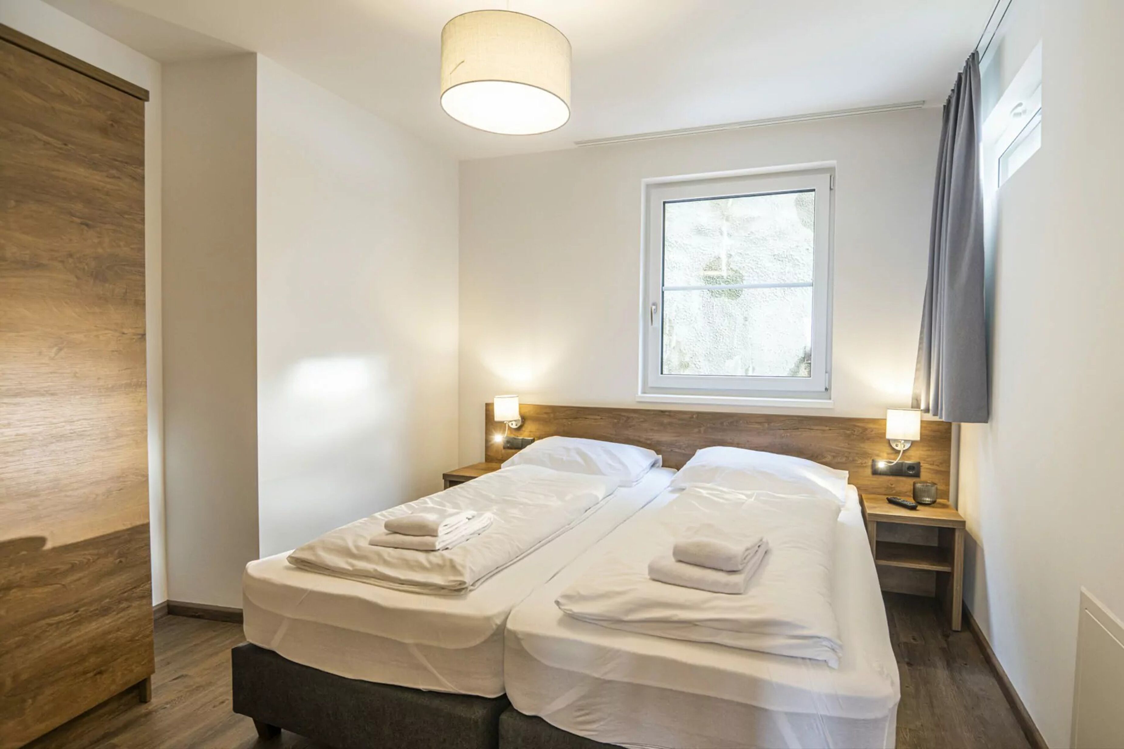 Une chambre d’hôtel avec deux lits simples, une armoire en bois, une fenêtre avec des rideaux et une lampe fixée au mur.