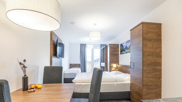 A compact hotel room with a wooden dining table, two gray upholstered chairs, a sofa, a bed with a white mattress, a wooden wardrobe, and a large window with curtains. - AlpenParks Hotel & Apartment Alpendorf (Sankt Johann im Pongau)