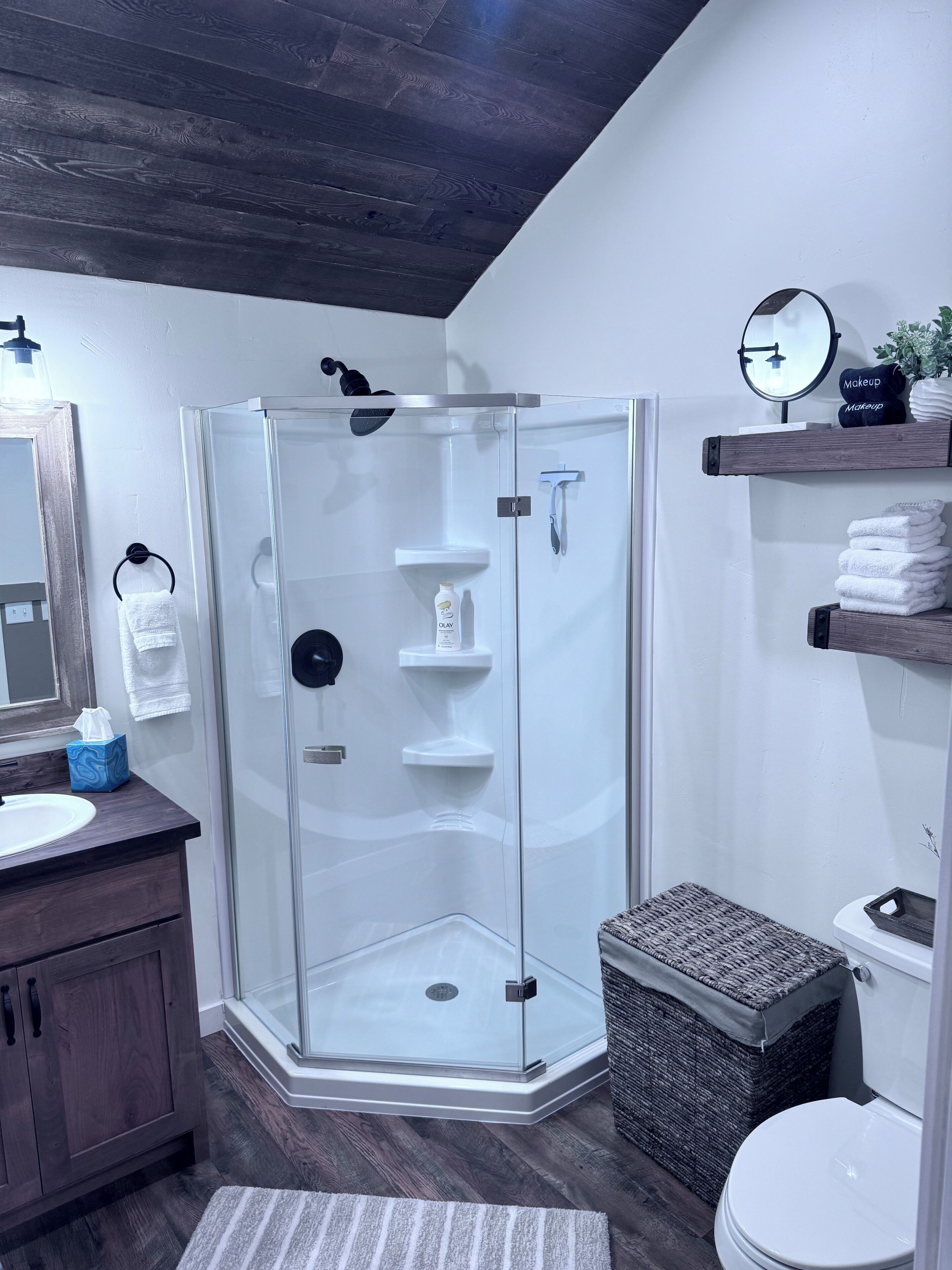 A modern bathroom with a glass shower enclosure, a wooden vanity, a round mirror, and a shelf with towels and toiletries.