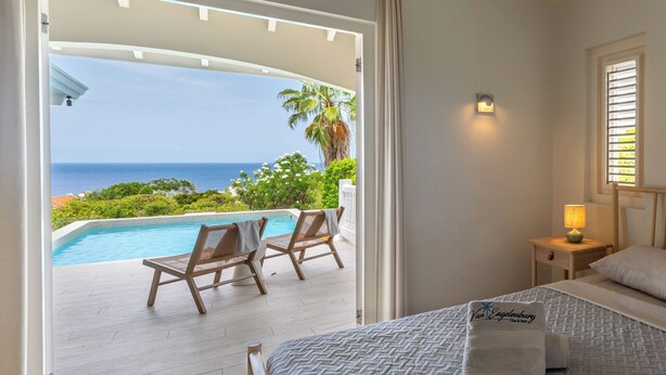 A hotel room with a bed, a nightstand, a lamp, a view of the ocean, and a pool area with chairs. - Villa Gone Coco - Coral Estate Resort (Sint Willibrordus)