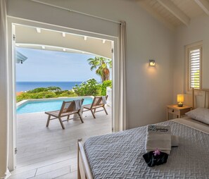 Ein Hotelzimmer mit Bett, Nachttisch, Lampe, Meerblick und einem Poolbereich mit Liegestühlen.