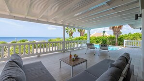 A covered outdoor seating area with a pool, white railings, and a view of the ocean. - Villa Gone Coco - Coral Estate Resort (Sint Willibrordus)