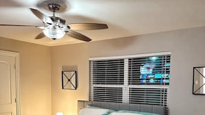 Un dormitorio con cama, ventilador de techo, ventana con persianas y mesita de noche con lámpara.