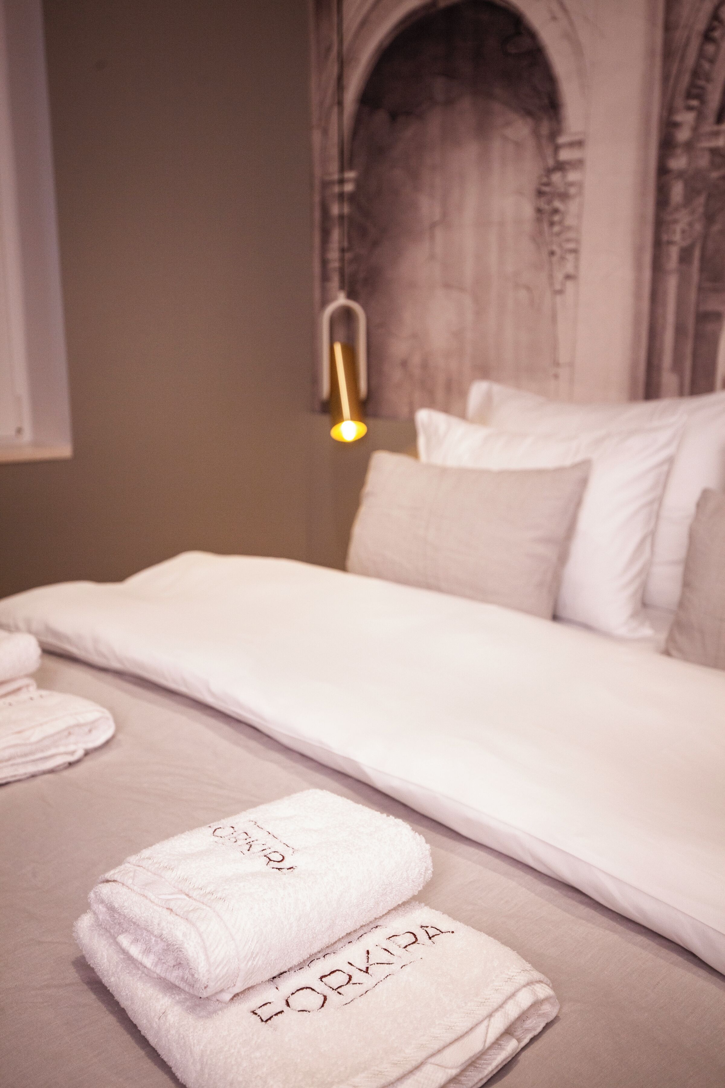 A neatly made bed with white linens and pillows, a bedside lamp, and two folded towels with text on them.