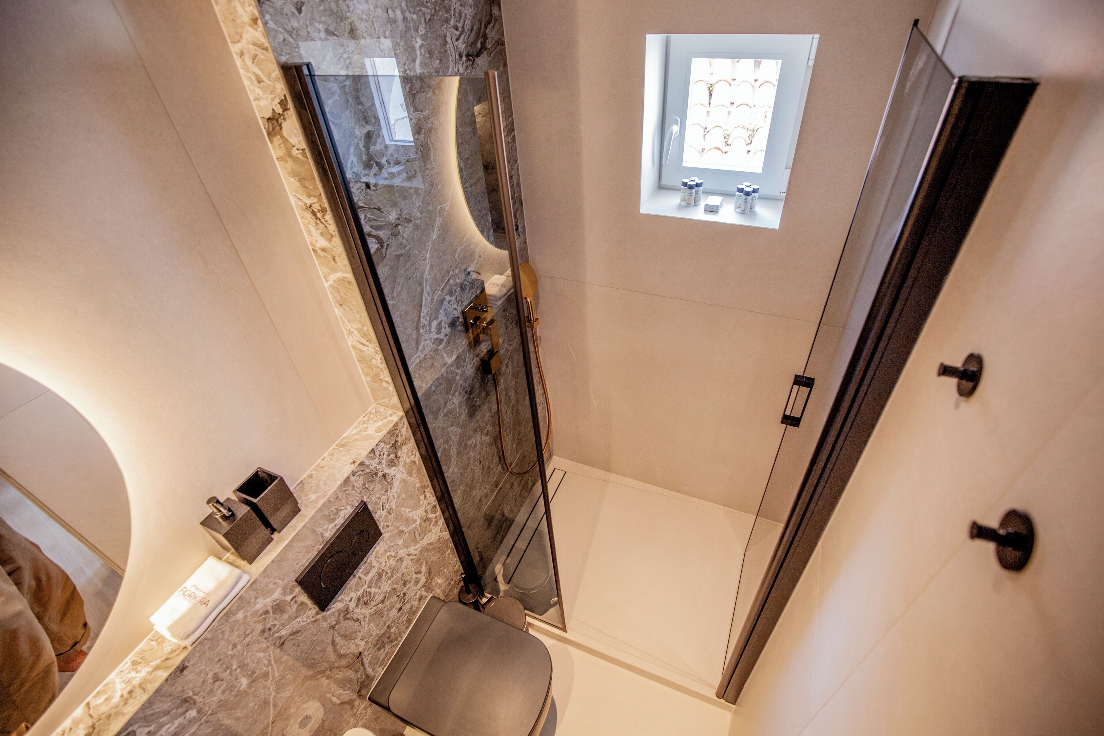 A modern bathroom with a glass-enclosed shower, marble countertop, and a small window providing natural light.