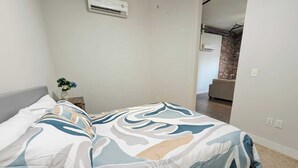 Un dormitori amb un llit, una tauleta de nit, un aparell d'aire condicionat i vista a una zona d'estar amb una paret de maó.