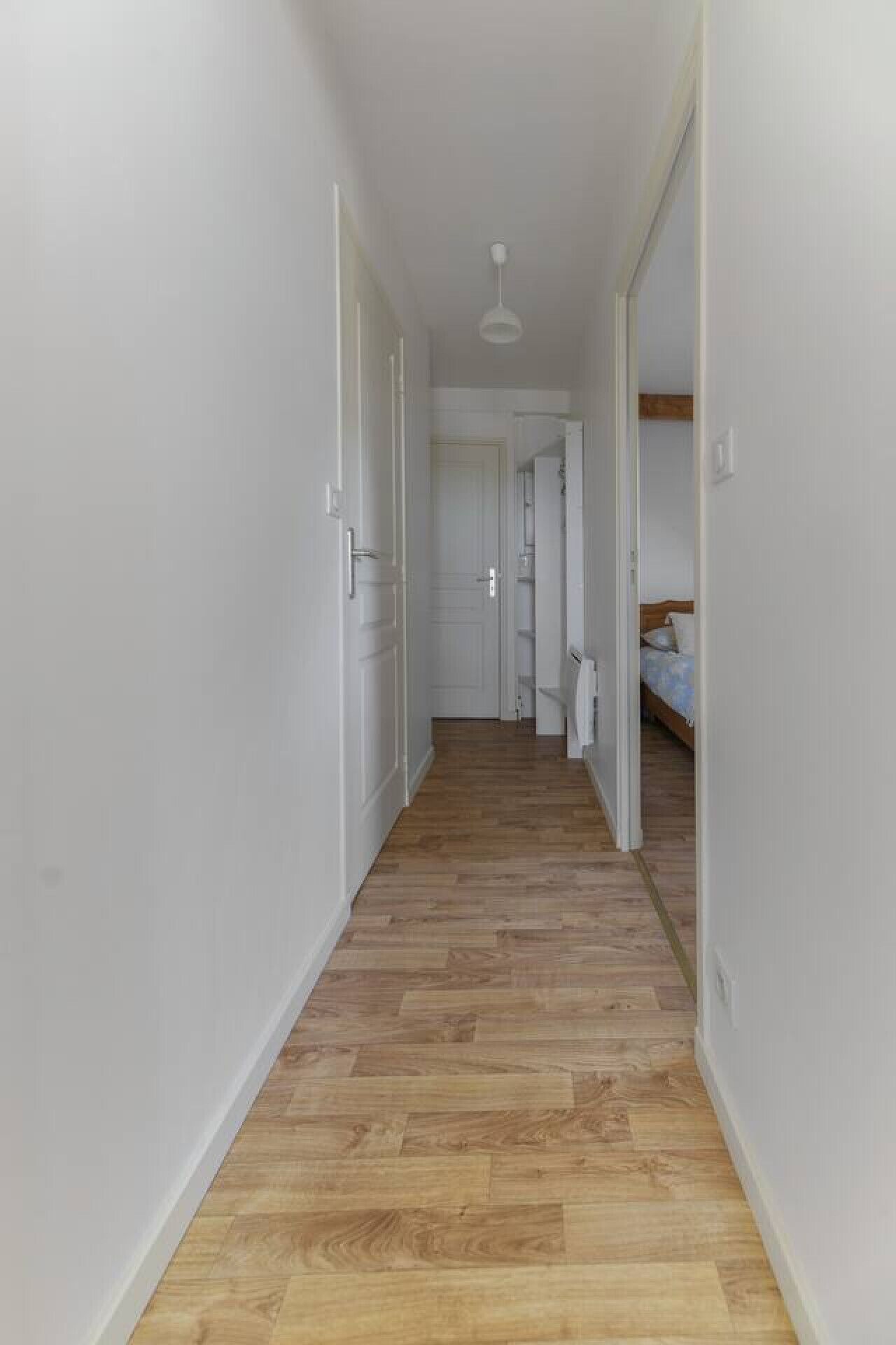 Un corridoio stretto con pavimento in legno e pareti bianche, che conduce a una camera da letto con un letto e un comodino.
