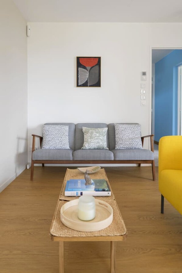 Een woonkamer met een grijze bank, gekleurde kussens, een houten bijzettafeltje met een boek en een glazen pot, en een gele stoel.