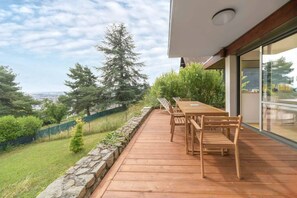 Een houten terras met een tafel en stoelen, met uitzicht op een grasveld met bomen en een stenen muur.