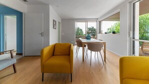 Una sala de estar moderna con un sofá amarillo, una mesa de comedor con sillas y grandes ventanas que dan a zonas verdes.