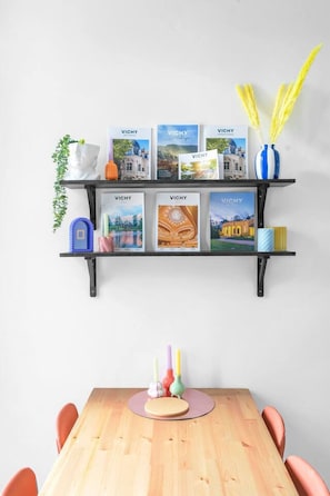 Ein Holztisch mit einer rosafarbenen Tischmatte, umgeben von orangefarbenen Stühlen. Darüber befinden sich zwei schwarze Regale mit verschiedenen Gegenständen, darunter Bücher, eine Vase mit gelben Blumen und ein weißer Brustbild.