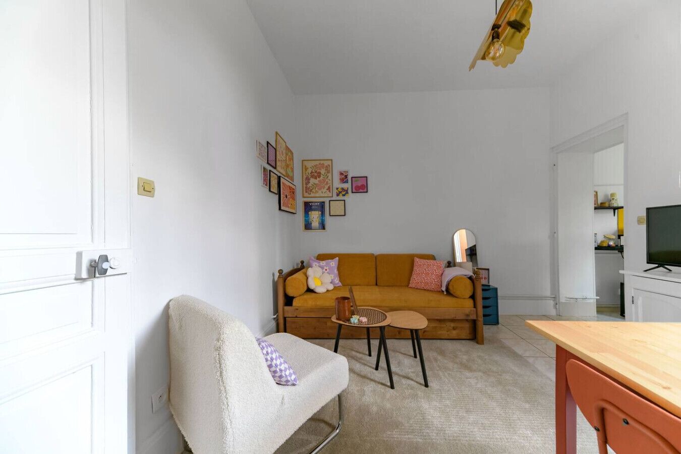 Una sala de estar con un sofá, una mesita redonda y una silla. Hay cuadros enmarcados en la pared y un televisor sobre un mueble.