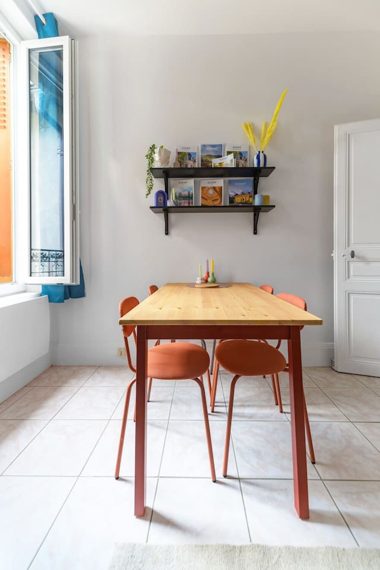 Un comedor con una mesa de madera y sillas naranjas, una estantería con libros y objetos decorativos, y una ventana con persianas.