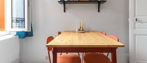 Un comedor con una mesa de madera y sillas naranjas, una estantería con libros y objetos decorativos, y una ventana con persianas.