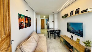 Een moderne woonkamer met een bank, een eethoek met stoelen, een aan de muur bevestigde televisie en een gang die naar een andere kamer leidt.