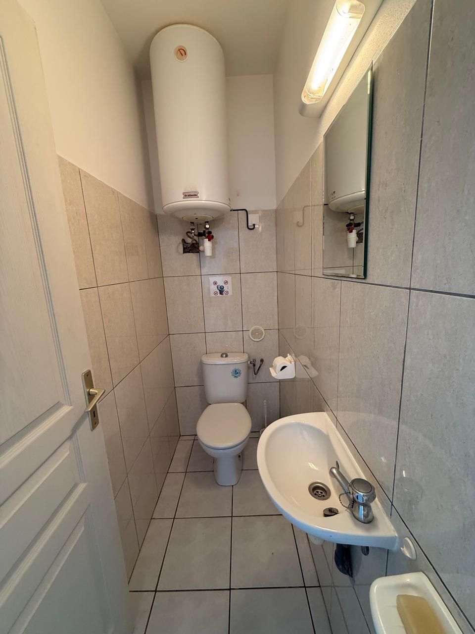 A small, modern bathroom with a toilet, sink, and bidet. It has tiled walls and floor, a mirror, and a ceiling light.