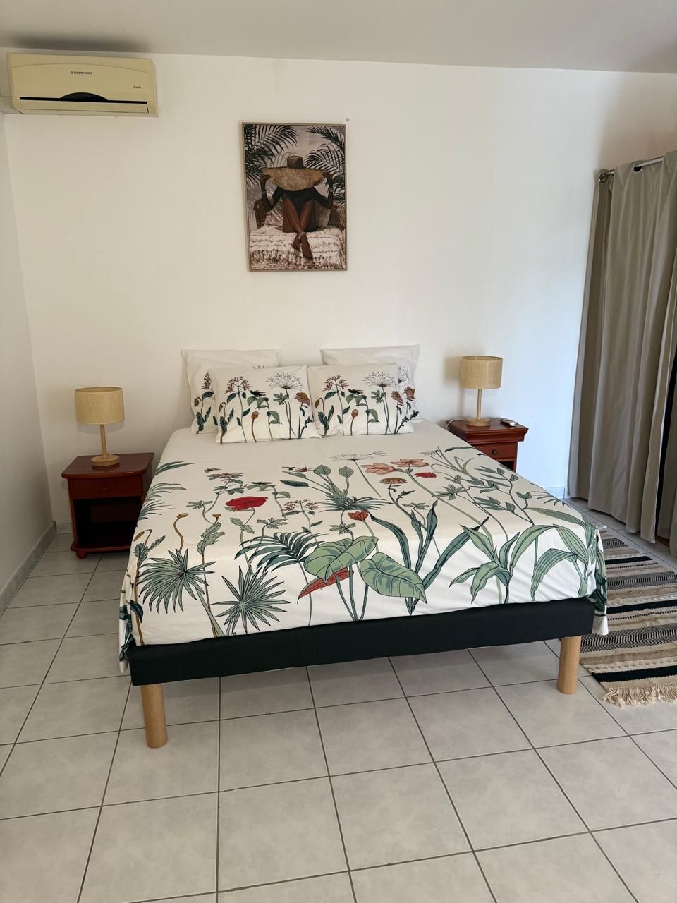 A bedroom with a bed, two nightstands, a wall-mounted air conditioner, a painting, and a window with curtains.