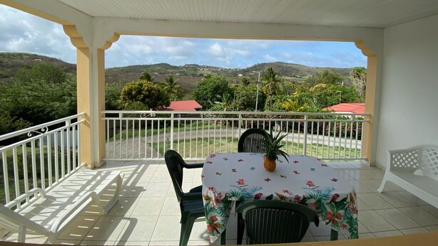 A covered outdoor area with a table and chairs, a white railing, and a view of hills and greenery.