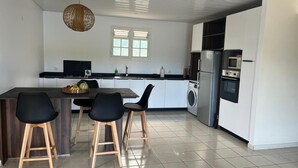 A modern kitchen with a dining area, featuring a central island with stools, a washing machine, and built-in appliances. - Alizé Cap (Sainte-Anne)