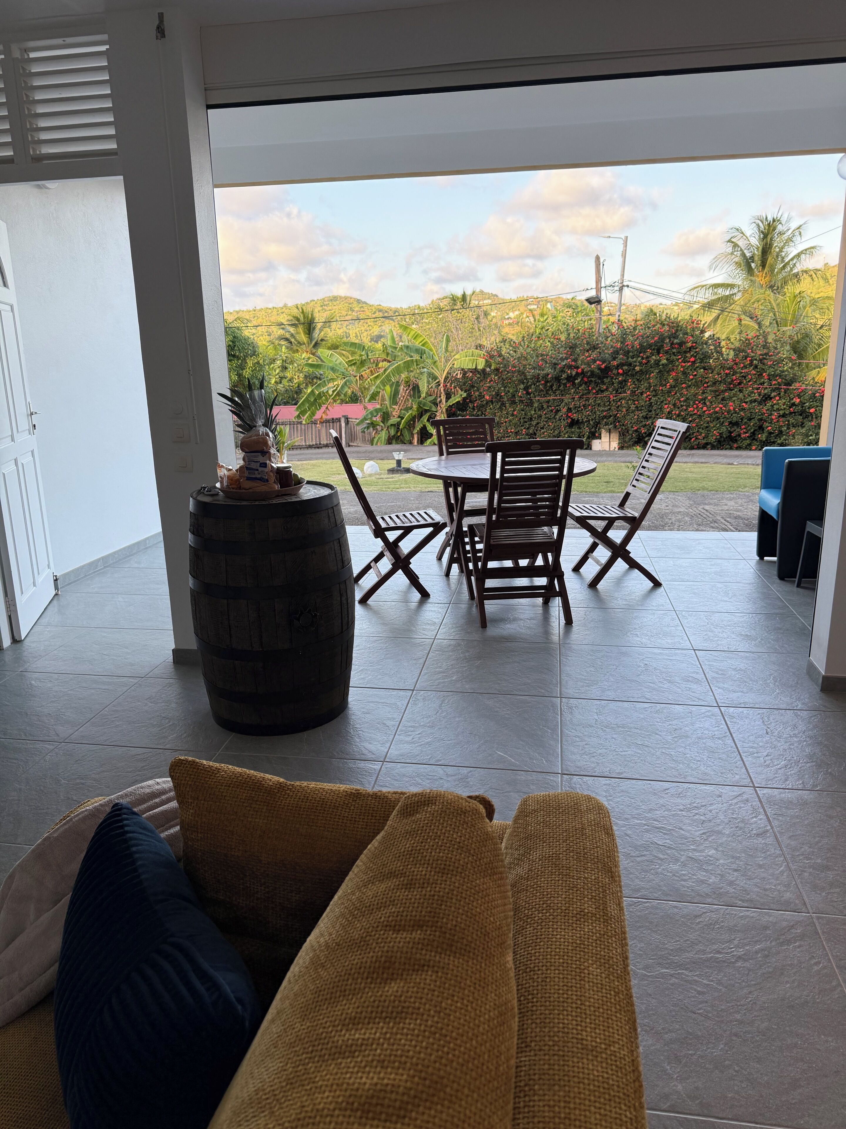 A patio area with a wicker sofa, a wooden table, and chairs. There is a barrel and a small decorative plant. The patio has a view of greenery and hills.