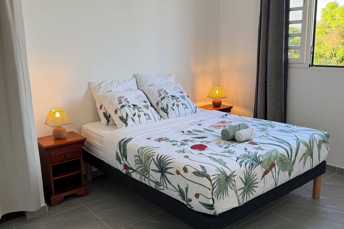 A bedroom with a bed, two bedside tables with lamps, a window with curtains, and an air conditioning unit on the wall.