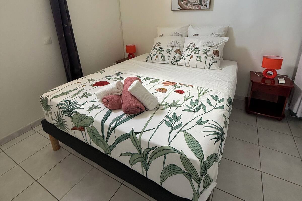 A neatly made bed with floral bedding, two bedside tables with lamps, a wall-mounted air conditioner, and a framed floral artwork above the bed.