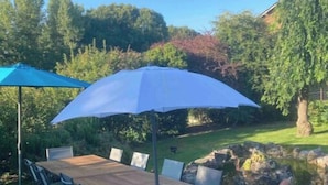 Un espacio al aire libre para comer con una mesa y sillas de madera, un paraguas azul y un jardín.