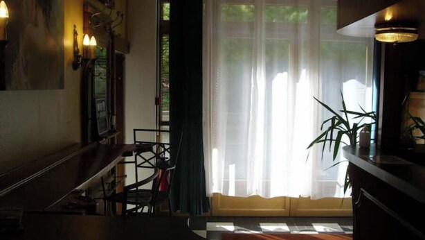 Una habitación con piso a cuadros, una mesa, sillas, una planta y vistas a través de una ventana con cortinas translúcidas.