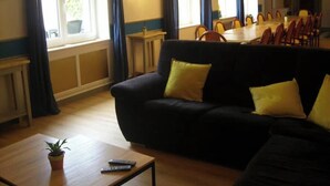 Una sala de estar con un sofá negro, cojines amarillos, una mesa de centro de madera con una planta y controles remotos, una zona de comedor con una mesa y sillas, y una lámpara fijada a la pared.