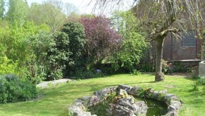 Un jardín con un estanque circular de piedra rodeado de césped y arbustos diversos.