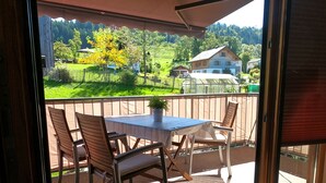 Uma varanda com mesa e cadeiras, um vaso de planta e vista para uma paisagem verde com casas.