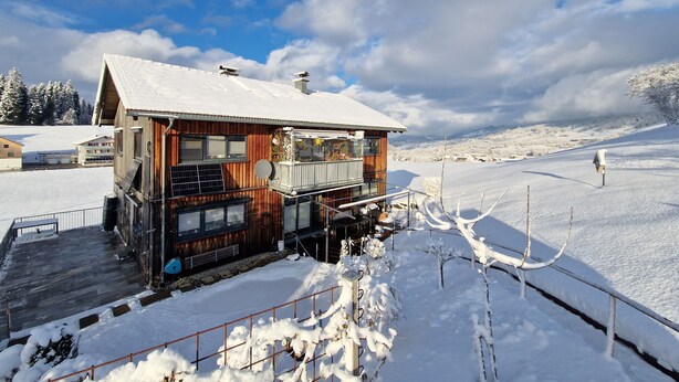 Uma cabana de madeira com varanda, rodeada por uma paisagem coberta de neve e montanhas ao longe.