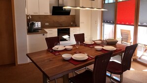 Uma sala de jantar com uma mesa de madeira posta para quatro pessoas, rodeada por cadeiras de vime. É visível uma cozinha ao fundo, com armários brancos e uma coifa preta.