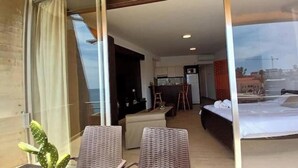 A hotel room with a bed, a small kitchenette, a seating area with a table and chairs, and a large window offering a view of the city. - Big Seaview studio, 24h electricity, beach + pool (Jabal Lubnan)
