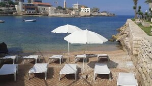 A beach with white umbrellas and lounge chairs, a stone wall, and a view of the sea and buildings in the background. - Big Seaview studio, 24h electricity, beach + pool (Jabal Lubnan)