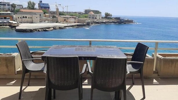 A terrace with wicker chairs and a table overlooking a coastal area with buildings and a pier. - Big Seaview studio, 24h electricity, beach + pool (Jabal Lubnan)