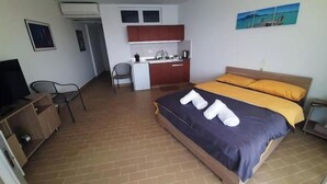 A compact hotel room with a bed, a desk, a chair, a small kitchenette, and a television.