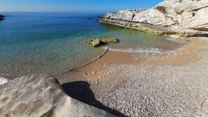 A coastal beach with clear water, pebbly shore, and rocky cliffs.