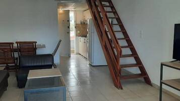 Un salon avec un canapé, une table basse, un téléviseur posé sur un meuble, une échelle en bois et, au fond, une cuisine.