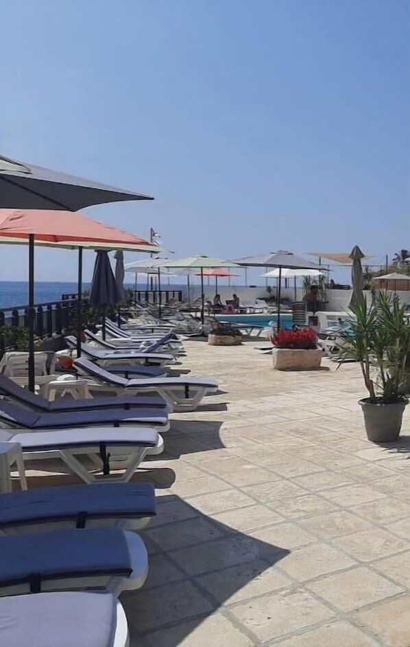 A beachside lounge area with sun loungers and umbrellas, a pool, and potted plants.