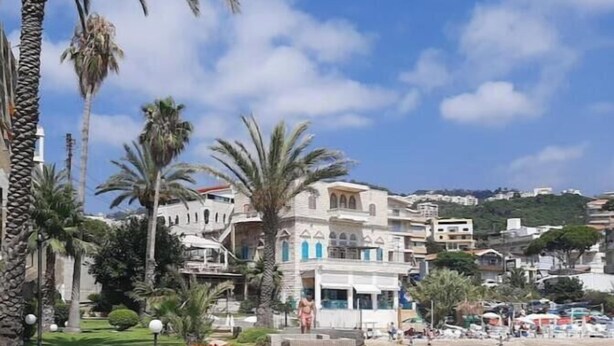 A coastal promenade with palm trees, a stone pathway, and a building with arched windows. - Family Duplex, beach+pool access (Jabal Lubnan)