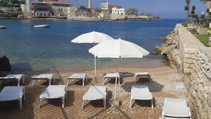 A beach with white lounge chairs and umbrellas, a stone wall, and a view of the sea and buildings in the background. - Family Duplex, beach+pool access (Jabal Lubnan)