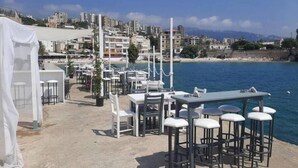 A seaside restaurant with outdoor seating, white tables and chairs, and a view of the sea and cityscape. - Family Duplex, beach+pool access (Jabal Lubnan)