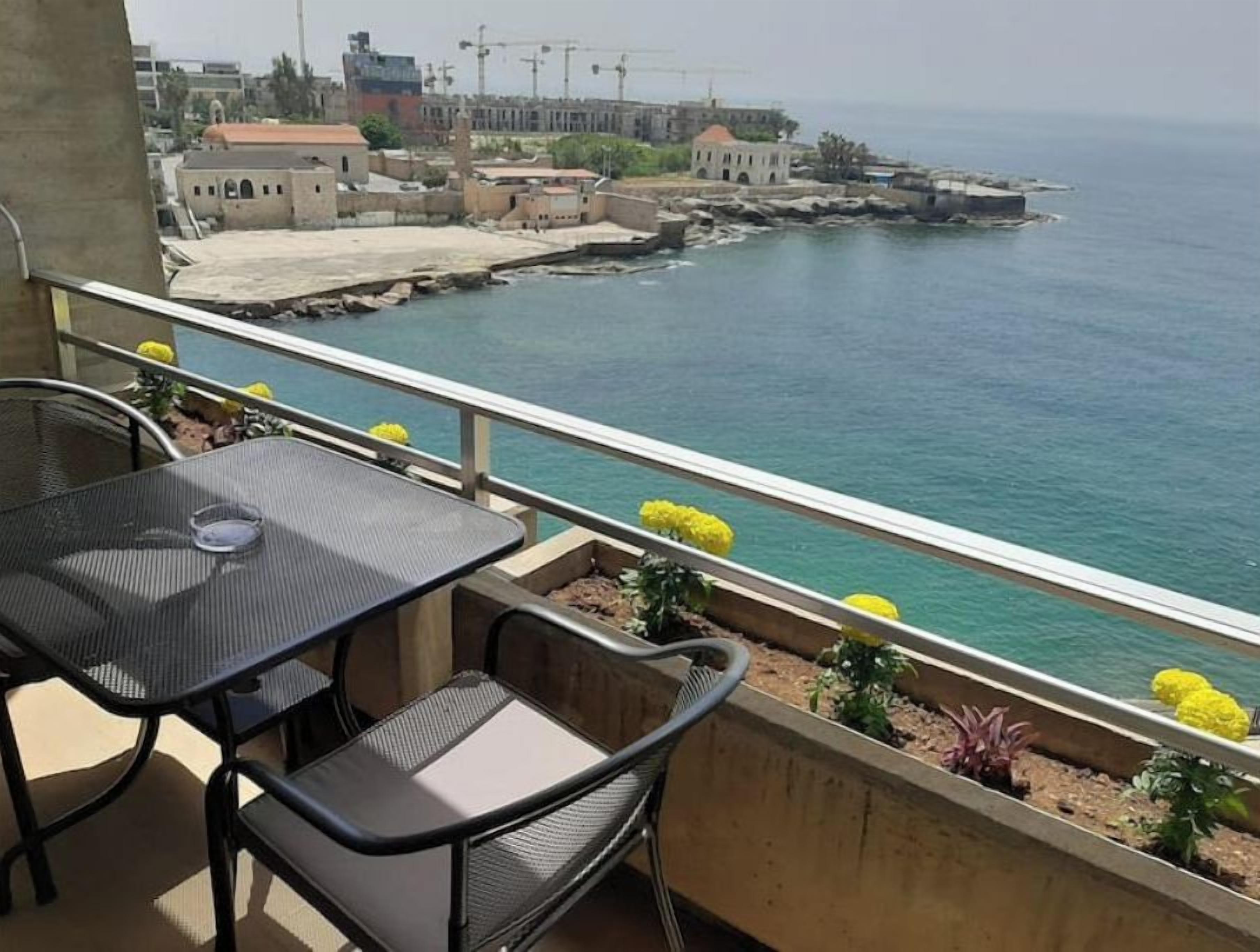 A balcony with a table and chairs overlooking a coastal area with buildings and a pier.